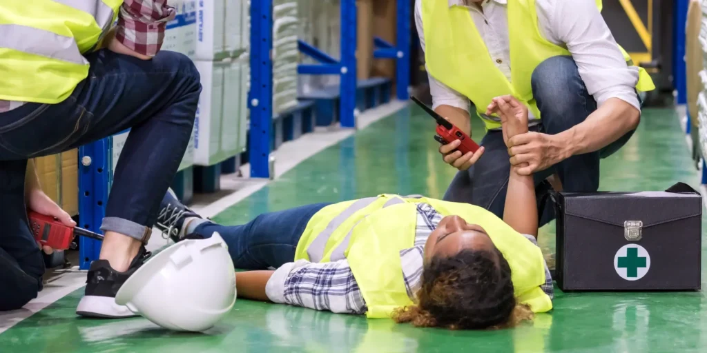 Ein Mann mit Sicherheitsweste liegt bewusstlos am Boden, zwei Männer geben Erste Hilfe, einer fühlt den Puls